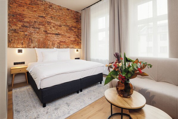 A hotel room with a brick wall, a bed, a sofa, a small table with a vase of flowers, and a window with curtains. - Nomadic Urban Premium Suites (Kraków)