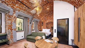A cozy room with a brick wall, a green sofa, a wooden table with chairs, and a bed with a white headboard. - Nomadic Urban Premium Suites (Kraków)