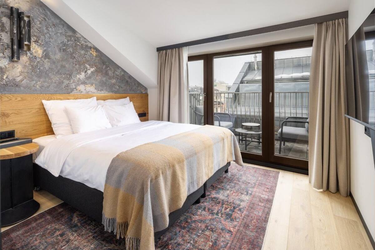 Ein modernes Hotelzimmer mit einem großen Bett, einem hölzernen Kopfteil, einem Nachttisch, einem gemusterten Teppich und einem Balkon mit Gartenmöbeln.