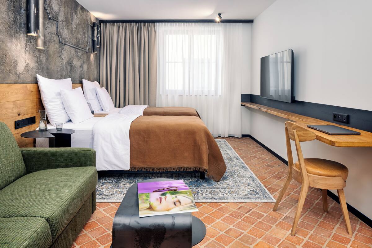 Ein Hotelzimmer mit Bett, Schreibtisch mit Laptop, grünem Sofa und Fernseher an der Wand.