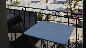 Ein Balkon mit Tisch und Stühlen, der auf einen Strand mit Menschen, Palmen und blauem Himmel blickt.