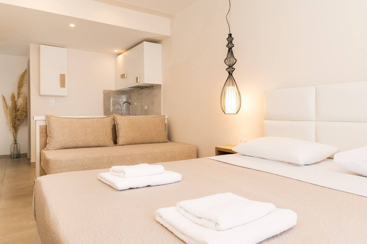 A modern hotel room with a large bed, a separate sofa, a kitchenette, and a hanging light fixture.