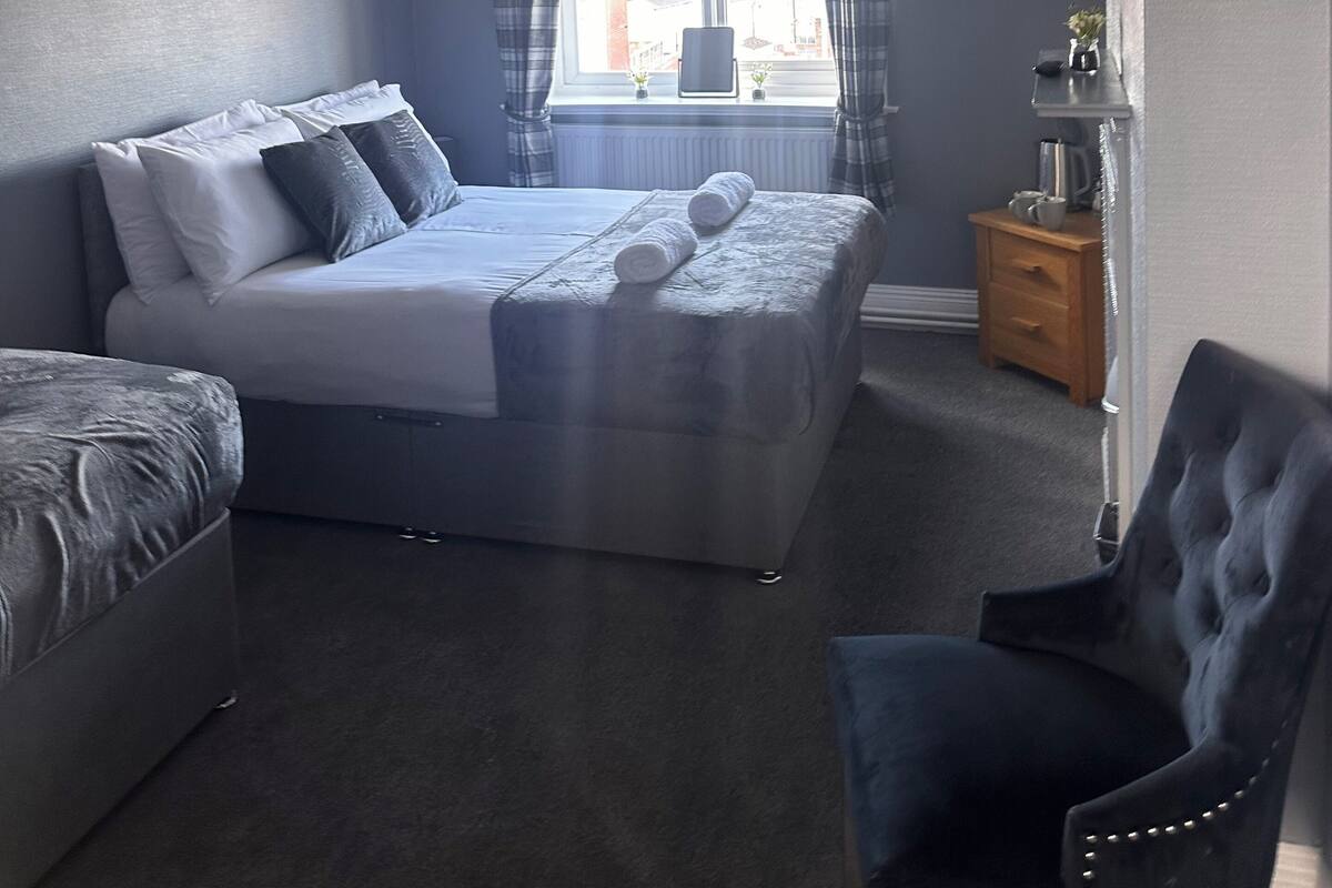 A bedroom with a bed, a chair, a bedside table, a window with curtains, a ceiling light, and a radiator.