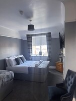 Un dormitorio con cama, una silla, mesita de noche, ventana con cortinas, luz de techo y radiador.