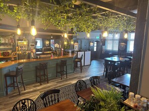 A bar area with a counter, stools, and hanging plants. There are tables and chairs in the dining area. - Sheridans (Wallasey)