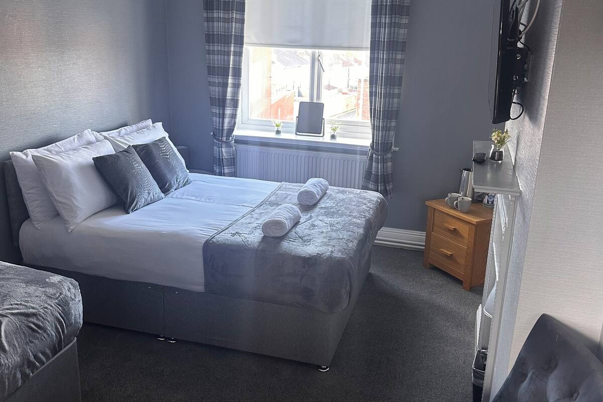 A bedroom with a bed, a chair, a bedside table, a window with curtains, and a wall-mounted TV.