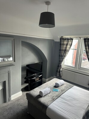 A bedroom with a bed, a television on a stand, a radiator, and a window with curtains.
