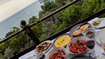 Una mesa con diversos platos y una copa de vino tinto, con vistas a un paisaje costero con vegetación y el mar.