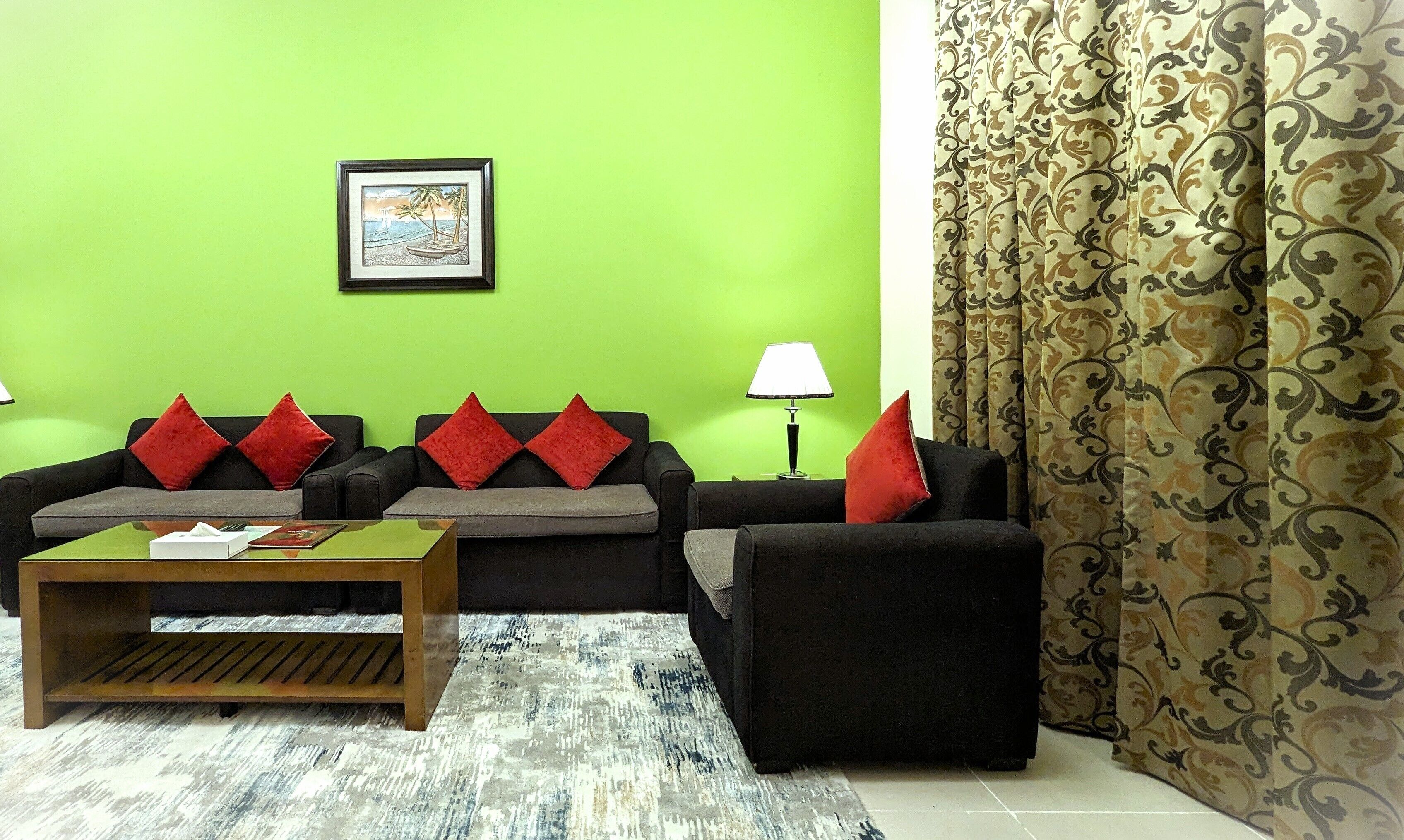 Un salón con una pared verde, un cuadro enmarcado, un sofá con cojines rojos, una mesa de centro y cortinas estampadas.
