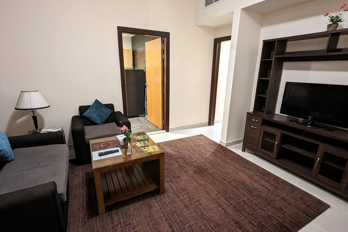 A living room with a sofa, coffee table, TV stand, and a door leading to a kitchen.