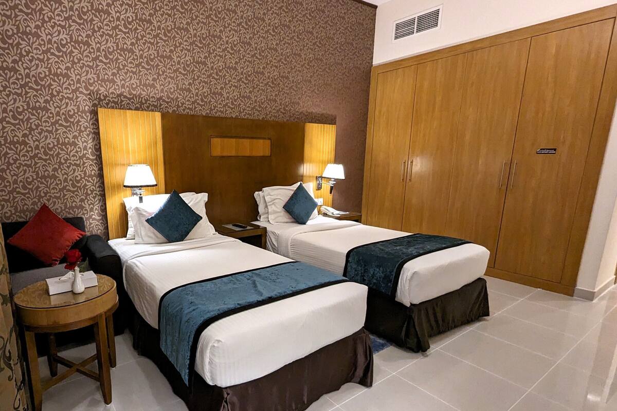 A hotel room with two beds, a wooden wardrobe, a nightstand with a lamp, and a red pillow.