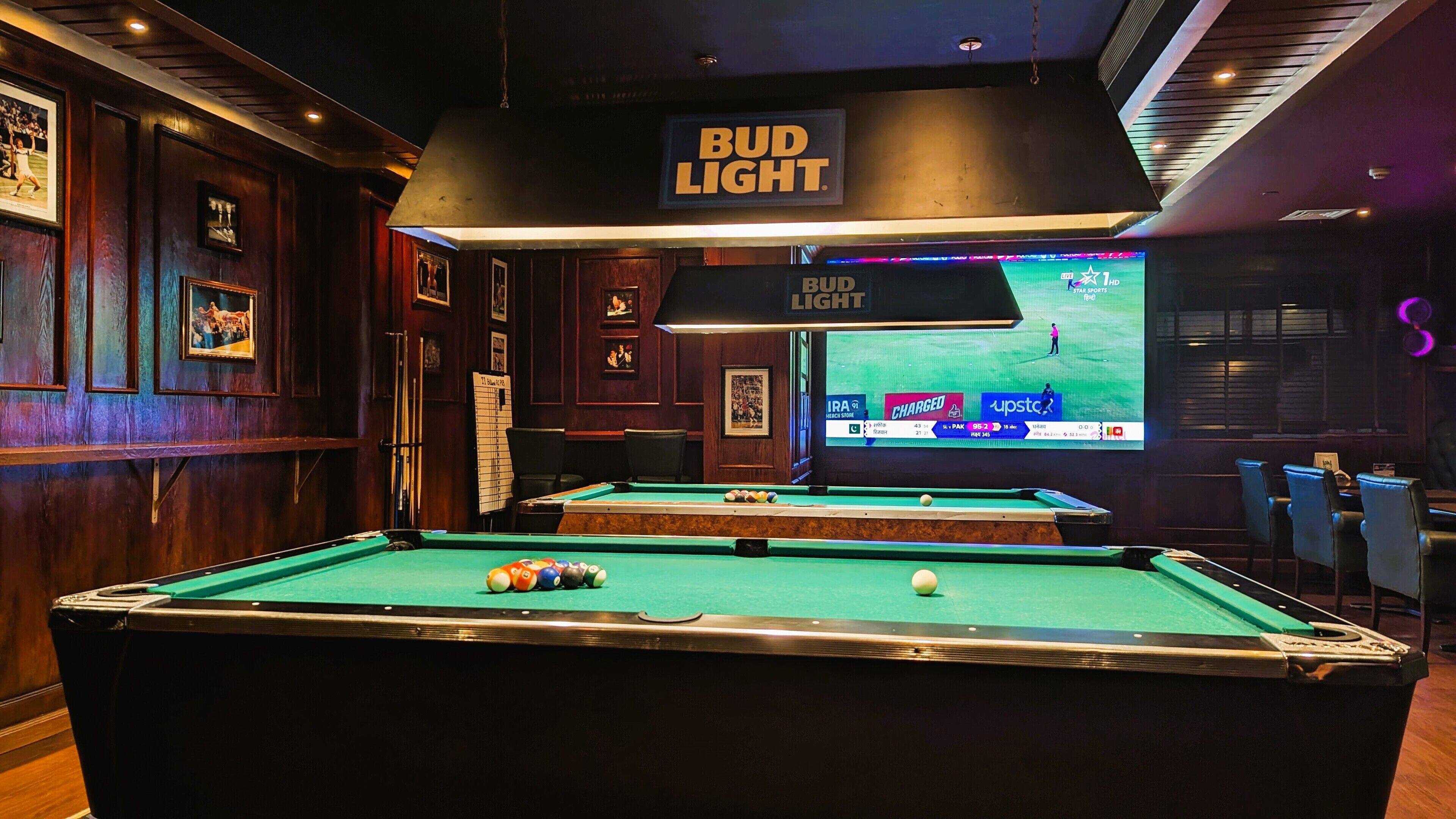 A pool table with a 'BUD LIGHT' sign above it, a television displaying a sports event, and a wooden wall with framed pictures.