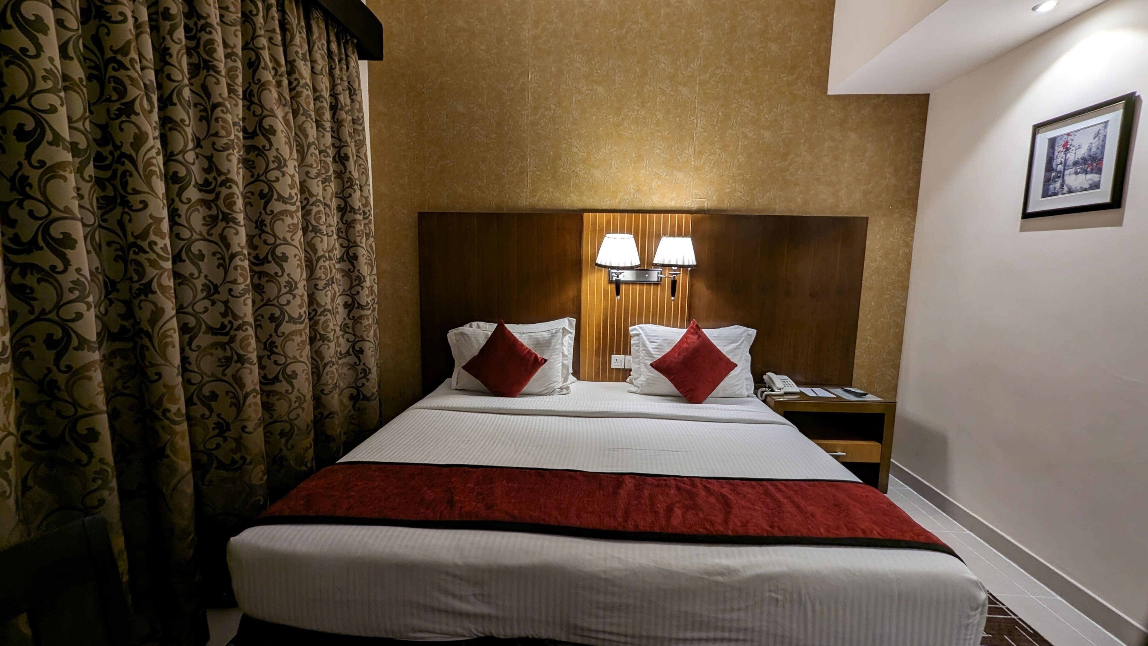 A hotel room with a large bed, two red pillows, a wooden headboard, a nightstand with a phone, and a wall-mounted lamp.