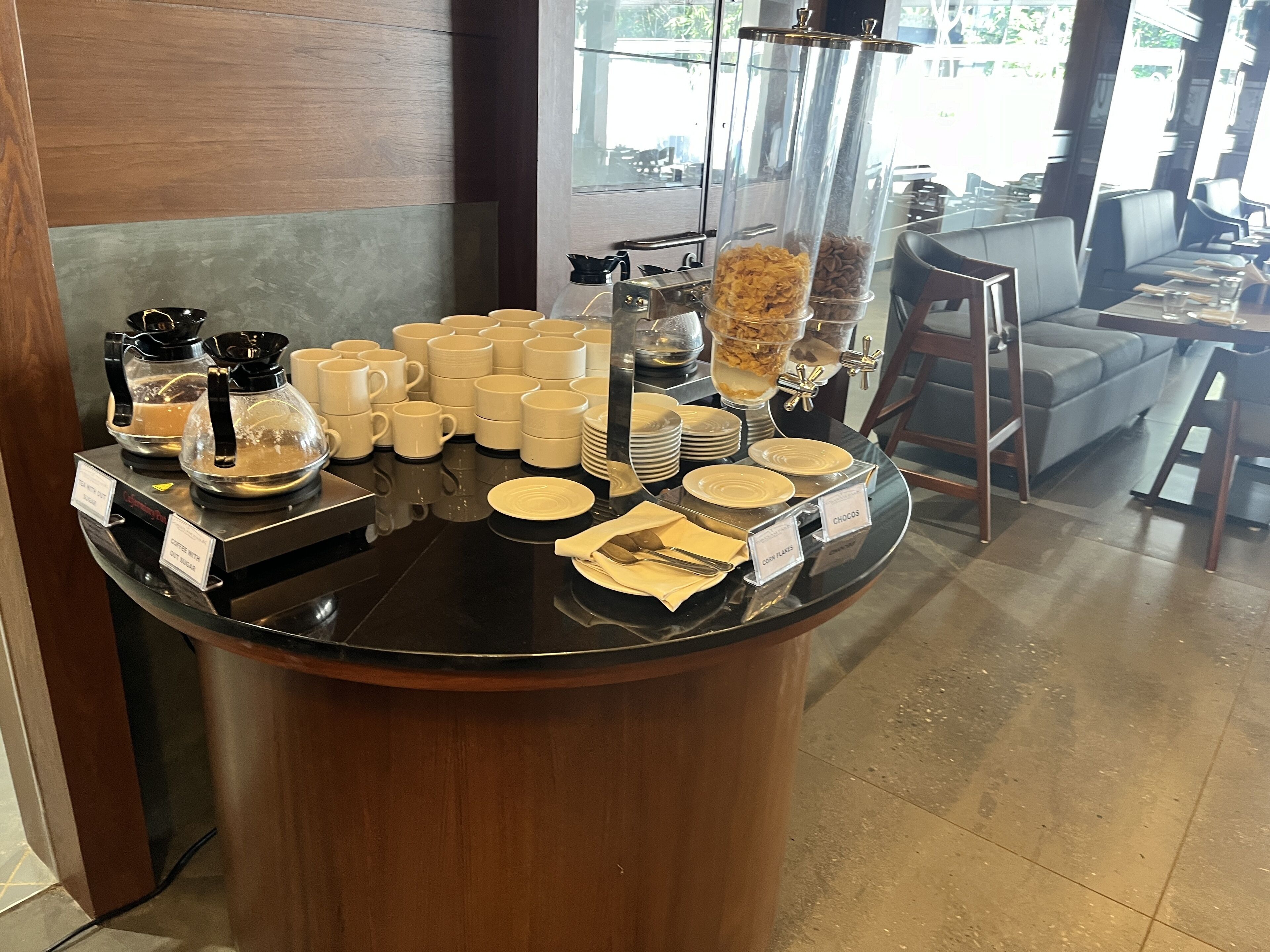 Una barra de buffet con cafeteras, tazas, platos y recipientes etiquetados para alimentos.