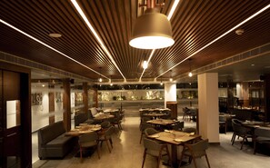 En moderne restaurant med træpaneler i loftet, hængende lamper og et centralt spiseområde med borde og stole.