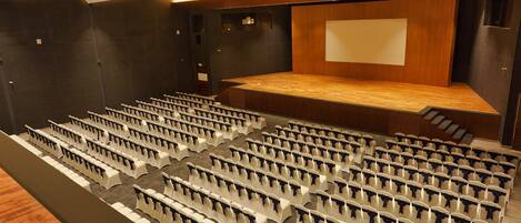 Un salón de conferencias con filas de sillas blancas, un escenario de madera y una pantalla grande.