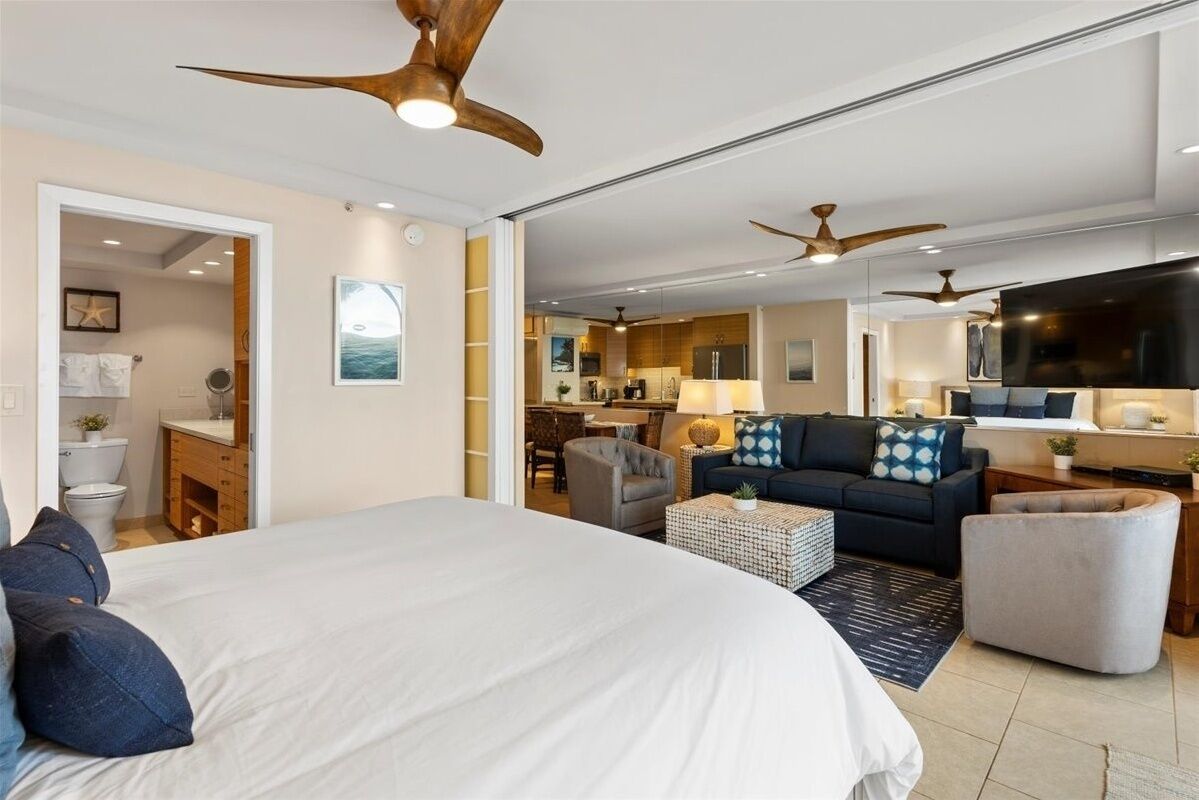 A modern hotel room with a large bed, a sofa, a dining area, and a kitchen in the background.