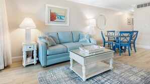 A living room with a blue sofa, a glass coffee table, a dining area with blue chairs, and a painting on the wall. - South Seas Bayside Villa 5226 1 Bedroom Condo by RedAwning (Captiva)