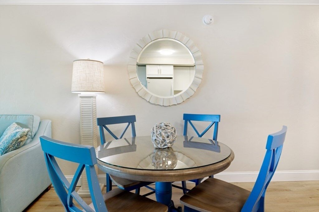 A dining area with a round glass table, blue chairs, a white lamp, and a wall-mounted mirror.