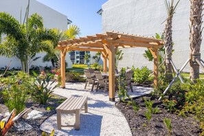 A wooden pergola with seating, surrounded by a garden with various plants and a gravel path.