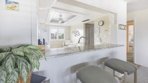 A kitchen with a central island, two bar stools, a ceiling fan, and a potted plant. - Loggerhead 424 2 Bedroom Condo by RedAwning (Sanibel)