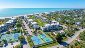 An aerial view of a coastal resort with multiple buildings, a tennis court, and a beach in the background. - Loggerhead 424 2 Bedroom Condo by RedAwning (Sanibel)