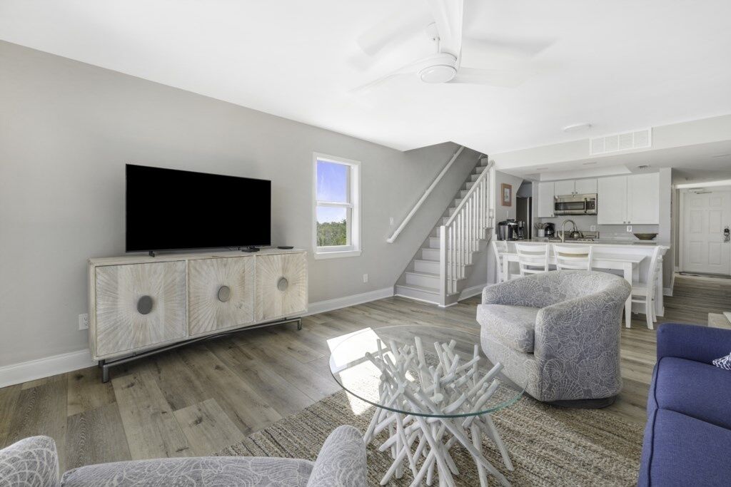 A modern living room with a flat-screen TV, a glass coffee table, and a staircase leading to a kitchen.
