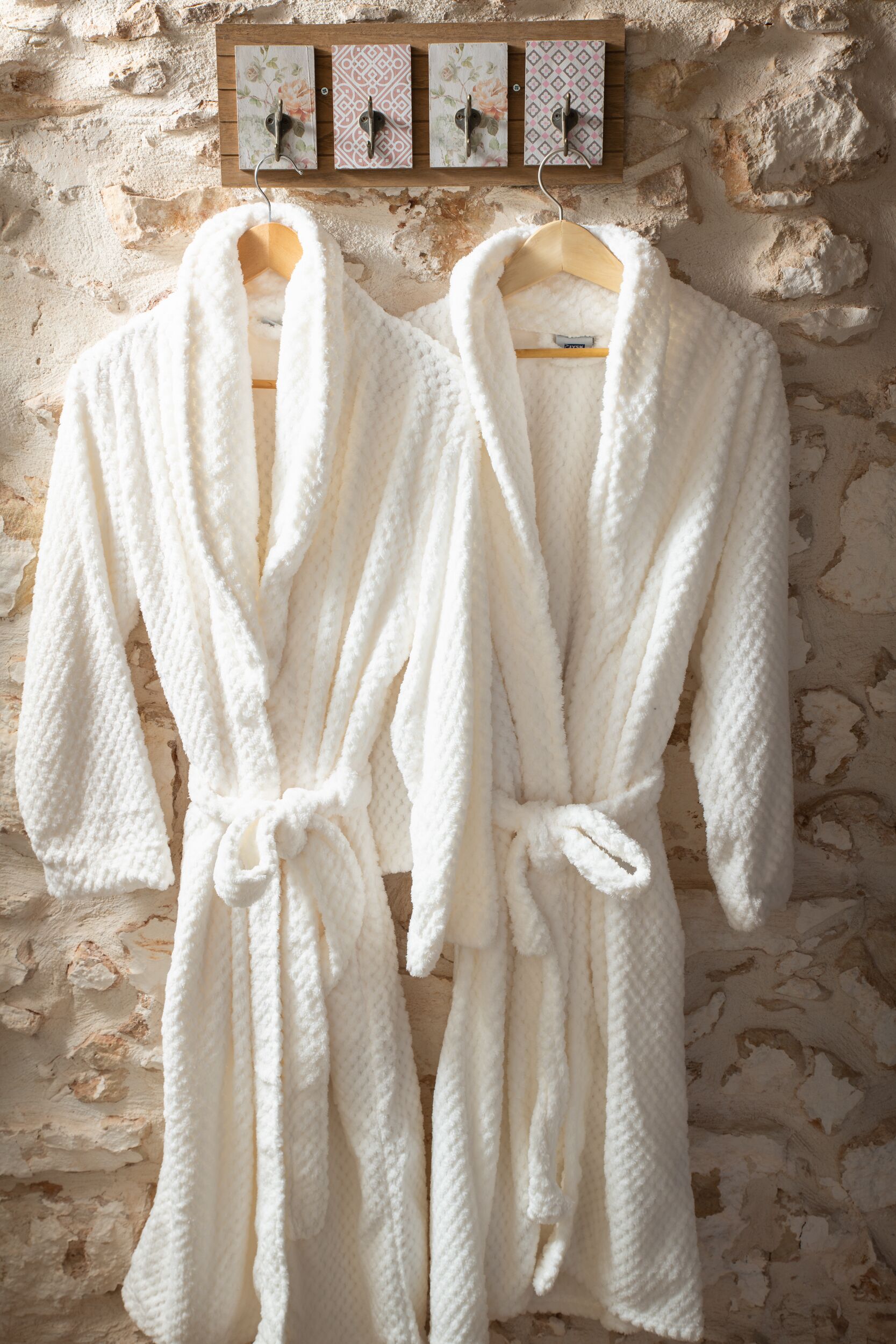 Two white bathrobes hanging on a wooden rack against a textured stone wall.