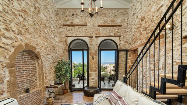A cozy room with stone walls, a wooden floor, a sofa, a table with a candle, and a view through arched windows. - Beit Petra Chania (Chania)