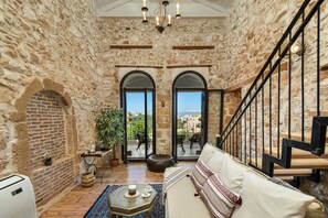 A cozy room with stone walls, a wooden floor, a sofa, a table with a candle, and a view through arched windows. - Beit Petra Chania (Chania)
