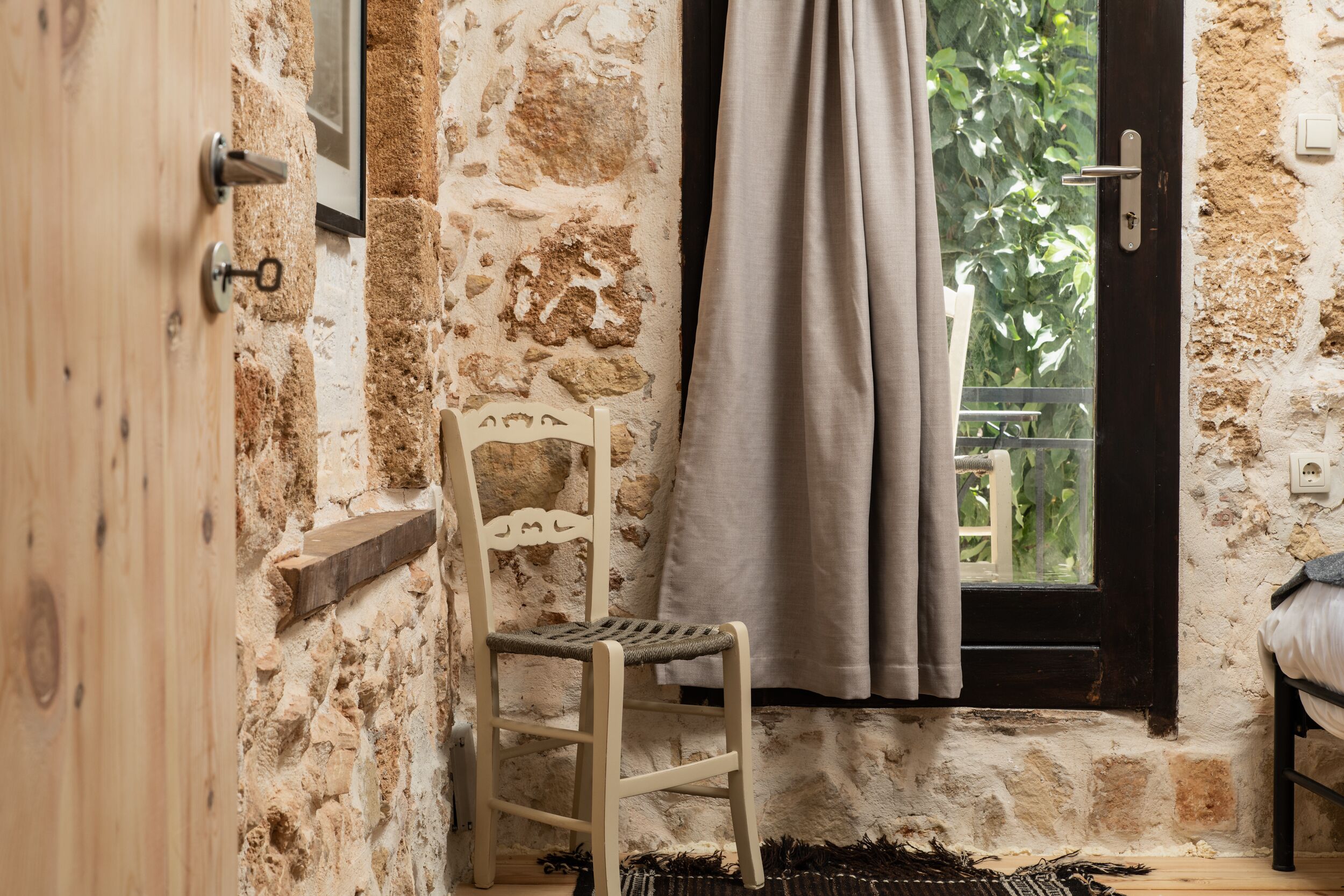 A rustic room with a stone wall, a wooden chair, a window with a curtain, and a bed.