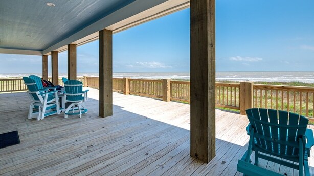 A covered deck with wooden flooring and a railing, featuring two green Adirondack chairs and a table.