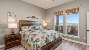 A bedroom with a large bed, two bedside tables, a ceiling fan, and a view of the outdoors through large windows.