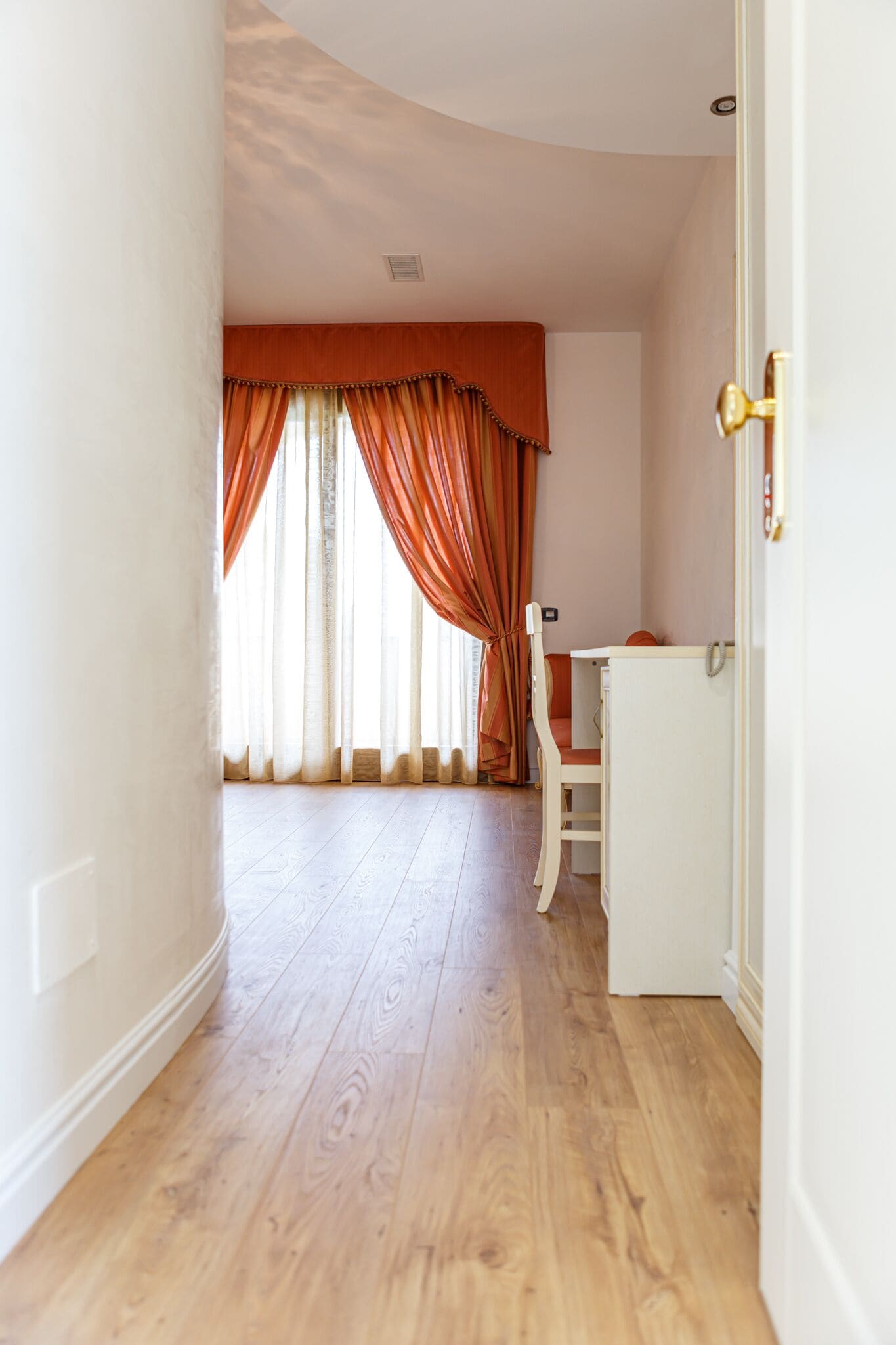 A hallway with wooden flooring, a white door with a gold handle, and a window with orange curtains.