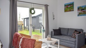 Un salon avec un canapé gris, un fauteuil en osier, une table basse blanche et une vue sur une terrasse en bois et la maison, visible à travers une porte vitrée.