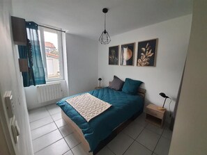 A bedroom with a bed, a nightstand, a lamp, a window with curtains, and wall art. - Appartement ABJ (Anneyron)