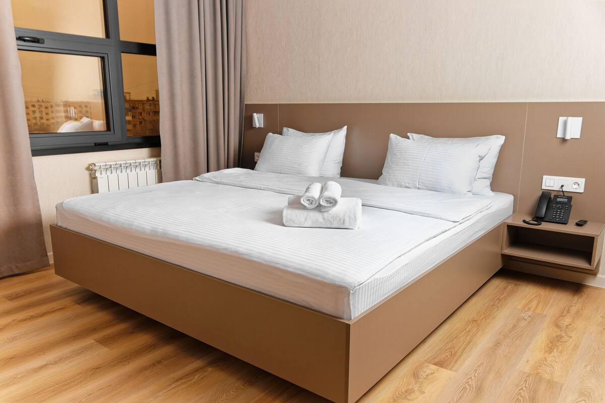 A modern hotel room with a large bed, a bedside table with a phone, and a view of buildings through the window.