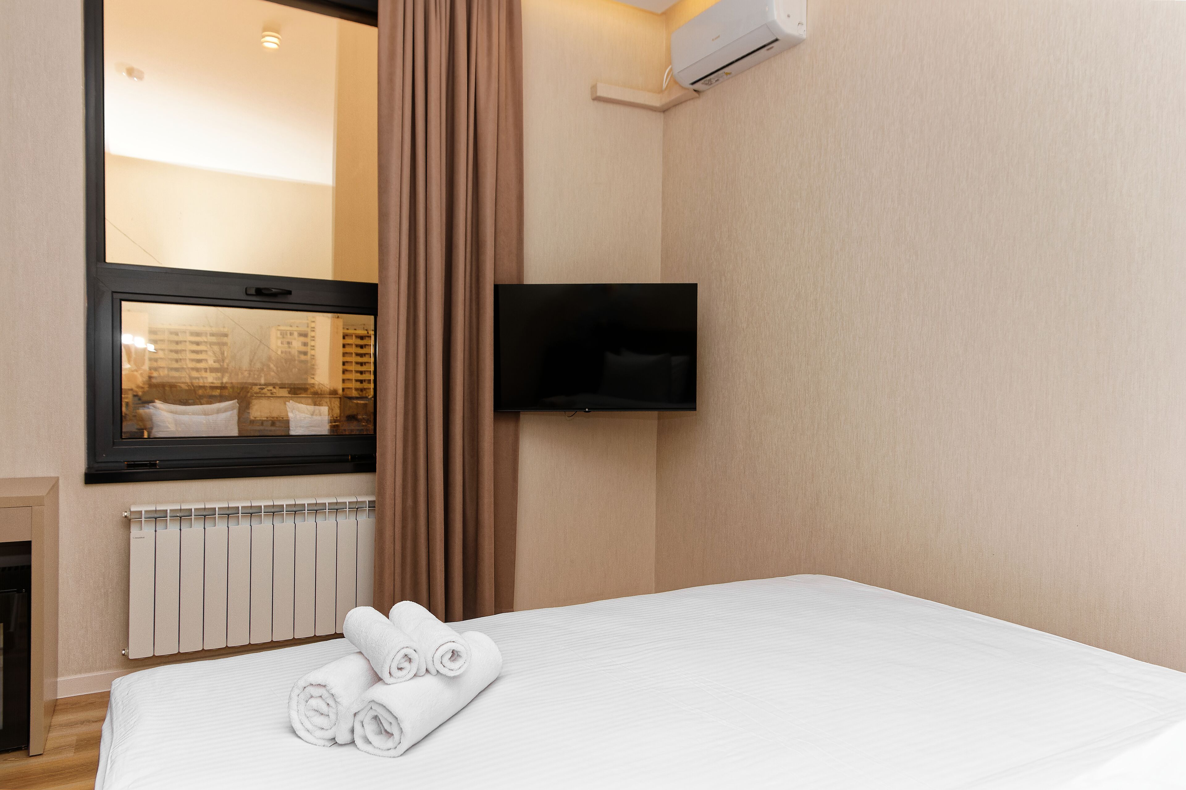A hotel room with a bed, a TV mounted on the wall, a window with curtains, and a radiator.