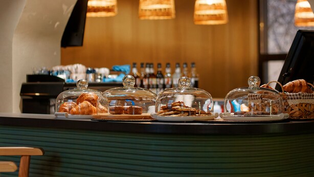 A bakery display with pastries under glass domes, a coffee machine, and a counter with a cash register. - Aston Hotel (Aktau)