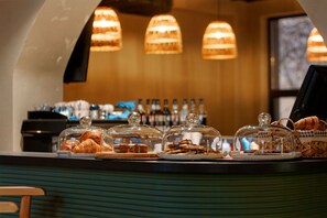 Una vitrina de pastelería con productos bajo campanas de cristal, una máquina de café y una barra con una caja registradora.