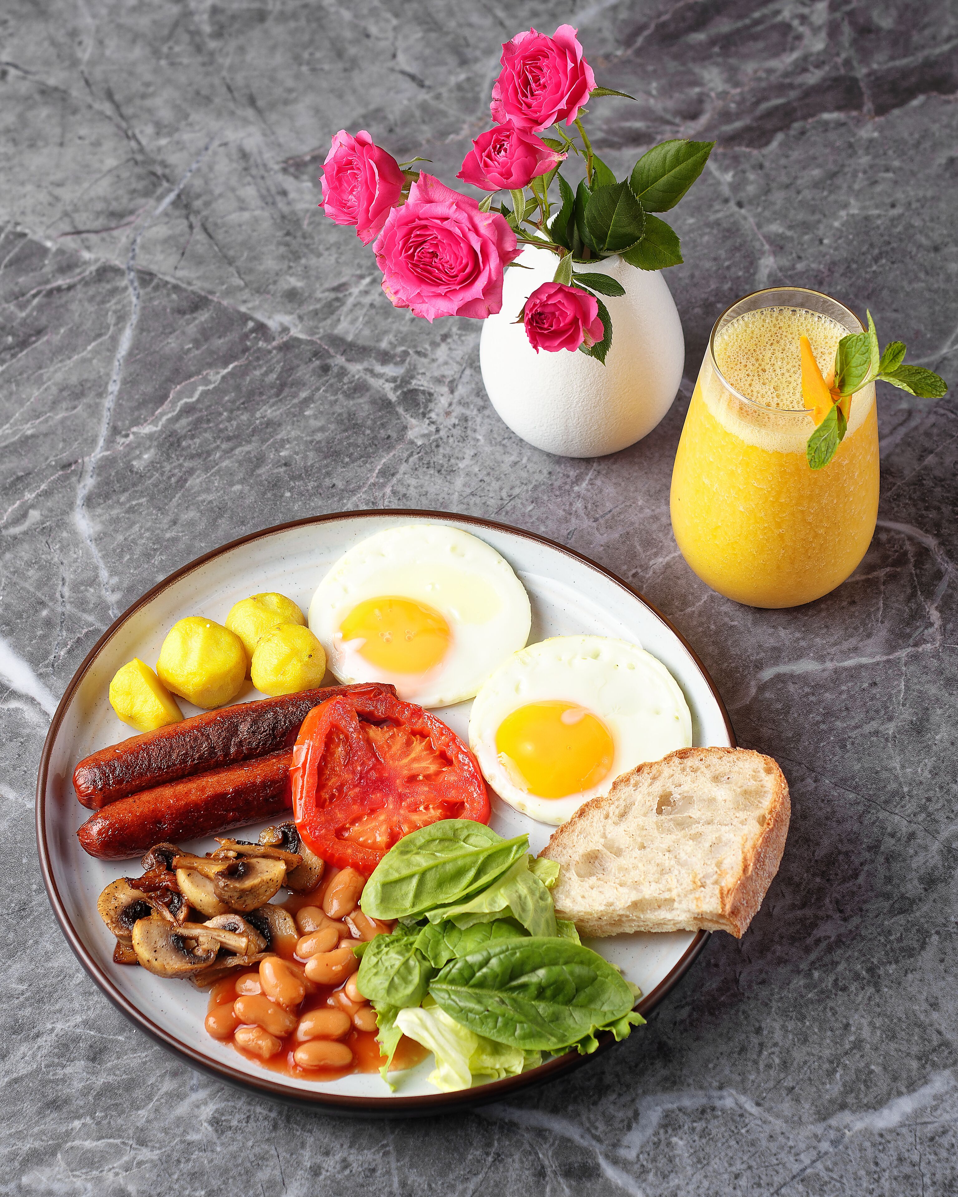 A breakfast plate with eggs, sausages, tomatoes, beans, mushrooms, spinach, and toast, along with a glass of orange juice and a vase of pink roses.
