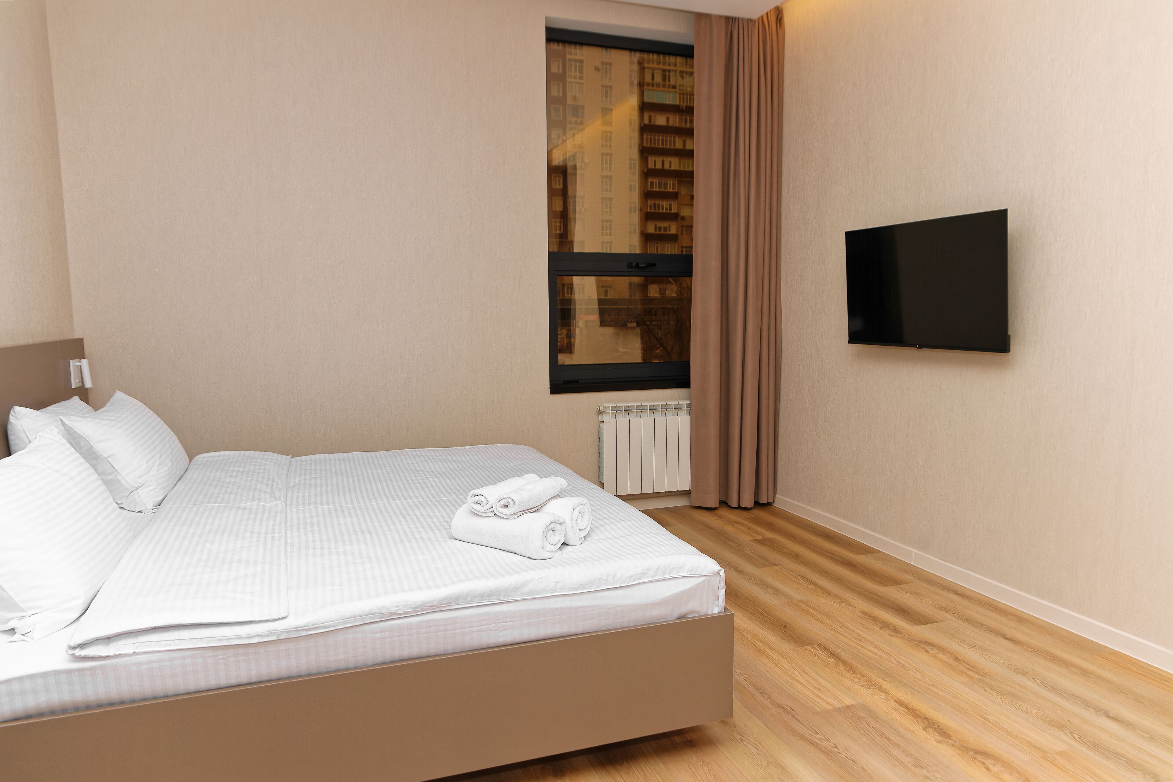 Un lit bien fait, avec du linge de lit blanc et des serviettes pliées, un téléviseur à écran plat fixé au mur et une fenêtre avec des rideaux.