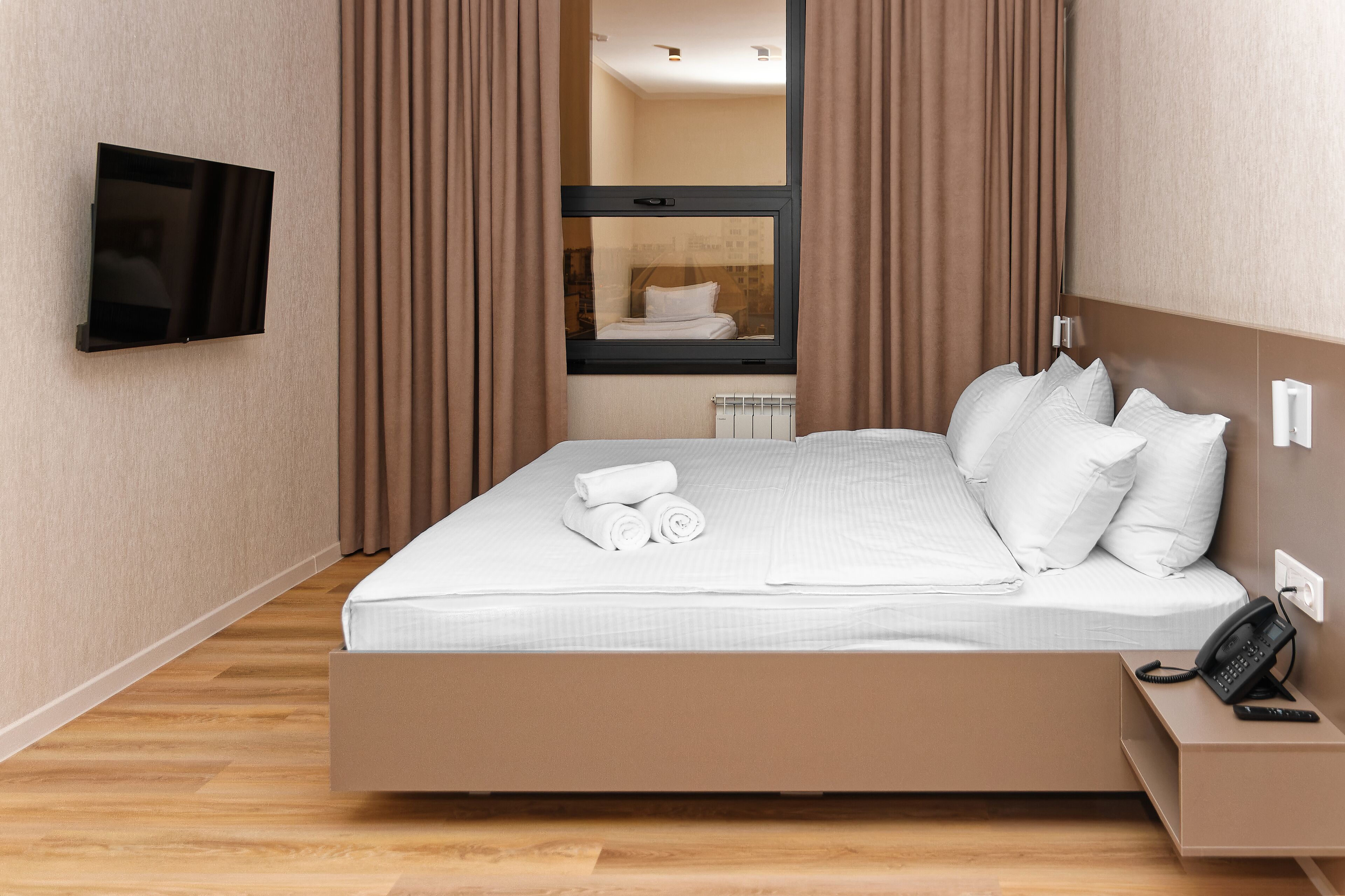 Quarto de hotel moderno com cama, televisão na parede, telefone ao lado da cama e janela com cortinas.