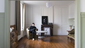 Ein Zimmer mit Holzboden, eine Person sitzt in einem Stuhl und liest ein Buch, ein kleiner runder Tisch, ein Kamin und ein gerahmtes Bild an der Wand.