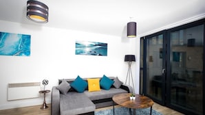A modern living room with a grey sofa, blue and yellow cushions, a round coffee table, and a large window with a view of a building. - Absolute Stays on Essex-chinatown-hippodrome-alexandra Theatre-bull Ring-the Rep (Birmingham)