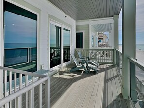Um deck coberto com vista para o oceano, com guarda-corpo, área de estar com cadeiras e porta de vidro que dá para um espaço ao ar livre.