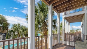 A covered outdoor area with a wooden deck, white columns, and a view of a swimming pool and palm trees. - Salty Beach | Shared Pool + Easy Beach Access! (Panama City Beach)
