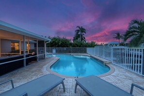 Ein Swimmingpool mit Liegestühlen, einer überdachten Sitzgelegenheit und Blick auf Palmen vor einem Sonnenuntergangshimmel.