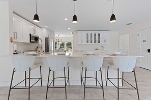 A modern kitchen with white cabinetry, a central island with bar stools, pendant lighting, and a view of the outdoors through large windows.