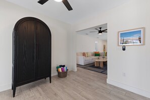 Ein modernes Wohnzimmer mit einem dunklen Holzschrank, einem Korb aus Weidengeflecht mit bunten Handtüchern, einem weißen Sofa mit Kissen, einem Couchtisch und einem Deckenventilator.